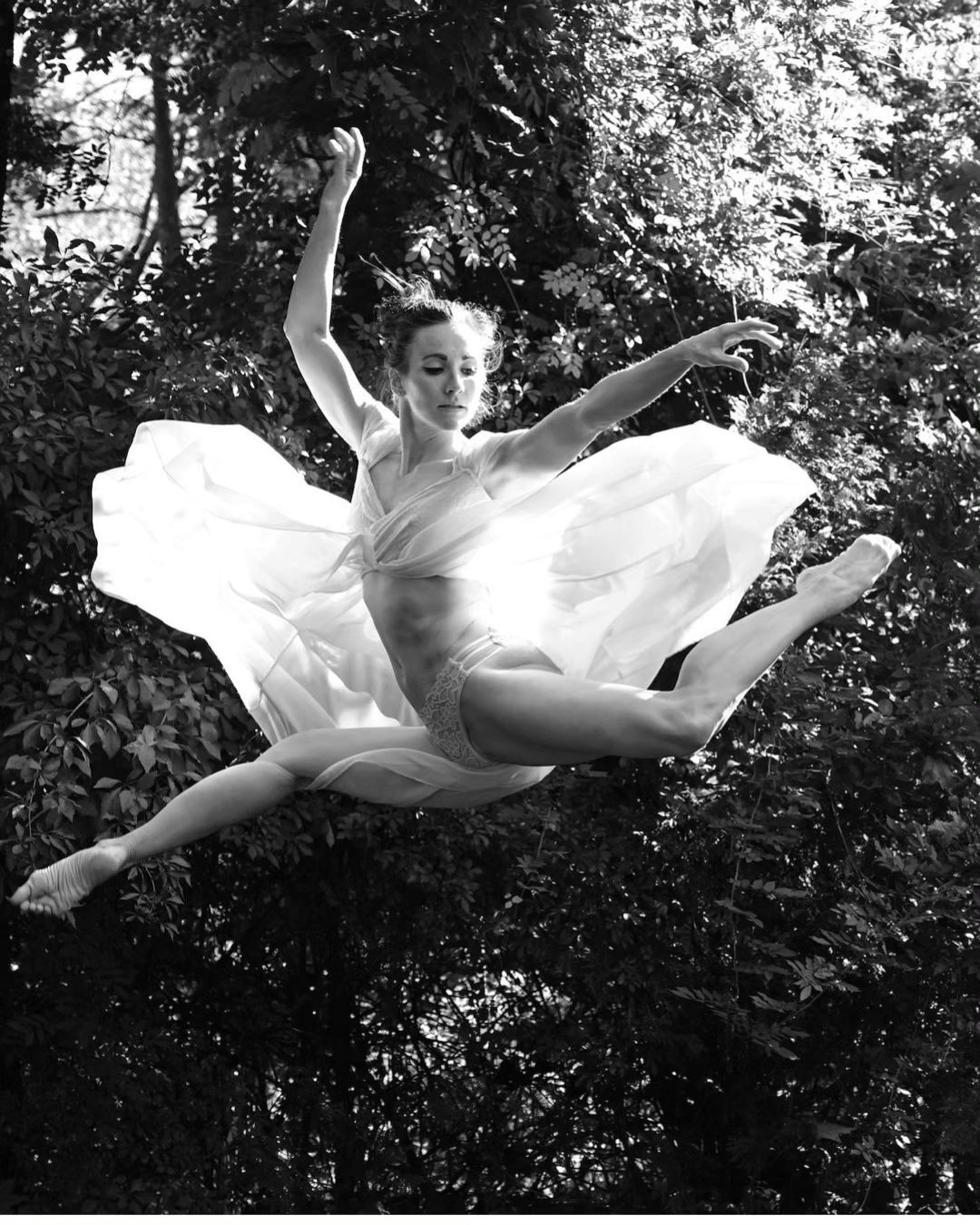 Woman in leotard and flowing fabric jumps against forest background, black and white. Artful shot of athleticism and nature.