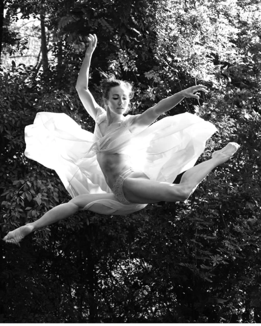 Woman in leotard and flowing fabric jumps against forest background, black and white. Artful shot of athleticism and nature.