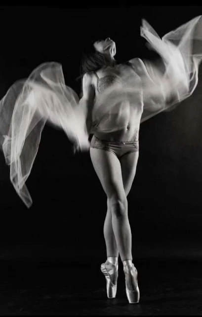 Monochrome photo of a woman in pointe shoes and dancewear, gracefully posing with arms raised in a dance studio, creating a stunning visual.