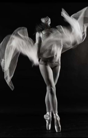 Monochrome photo of a woman in pointe shoes and dancewear, gracefully posing with arms raised in a dance studio, creating a stunning visual.
