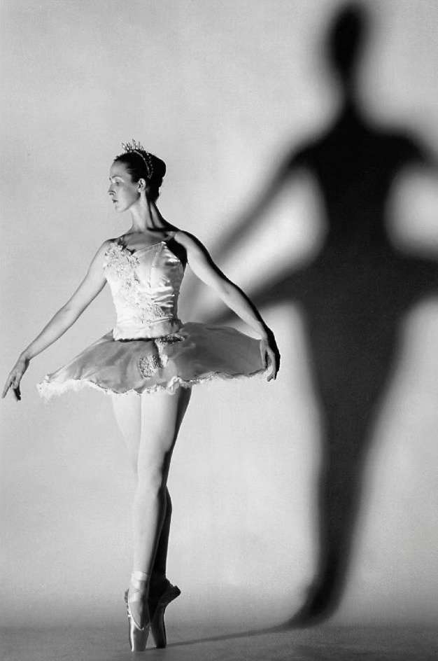 Elegant ballerina poses gracefully in pointe shoes and tutu, casting a dramatic shadow against a soft grey backdrop.