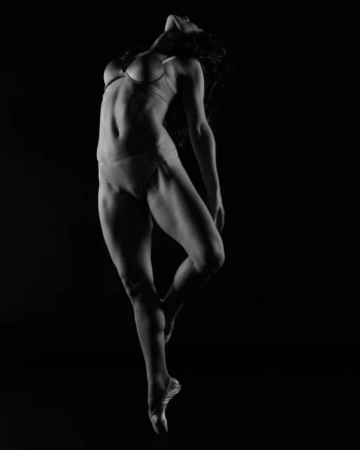 Artistic black and white studio portrait of an adult female dancer in a bra and briefs, leaping gracefully against a dark background.
