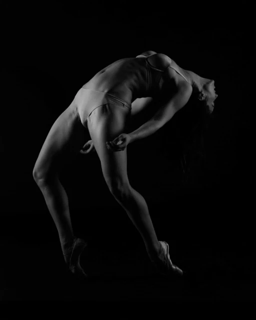 Monochrome portrait of a fit adult woman arching backwards in a contorted dance pose wearing ballet shoes against a dark background.
