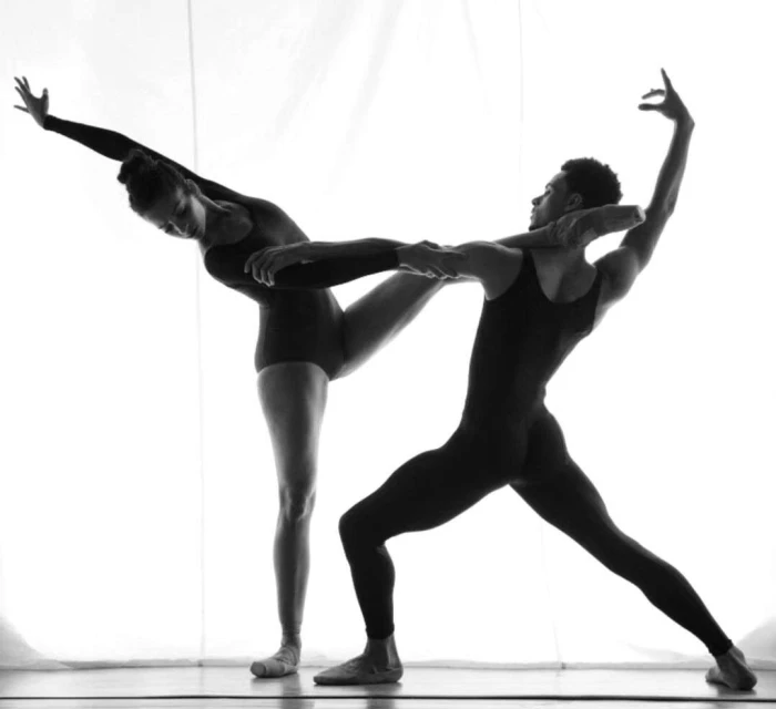 Two dancers, an adult man and an adult woman, pose in a studio with arms intertwined. They are dressed in black leotards.