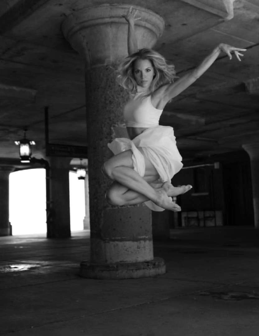 Adult woman mid-air leaps barefoot in dance pose near concrete column. She wears a cropped top and flowing skirt against a dark backdrop.