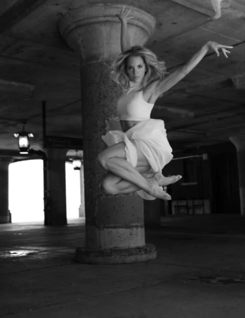 Adult woman mid-air leaps barefoot in dance pose near concrete column.  She wears a cropped top and flowing skirt against a dark backdrop.