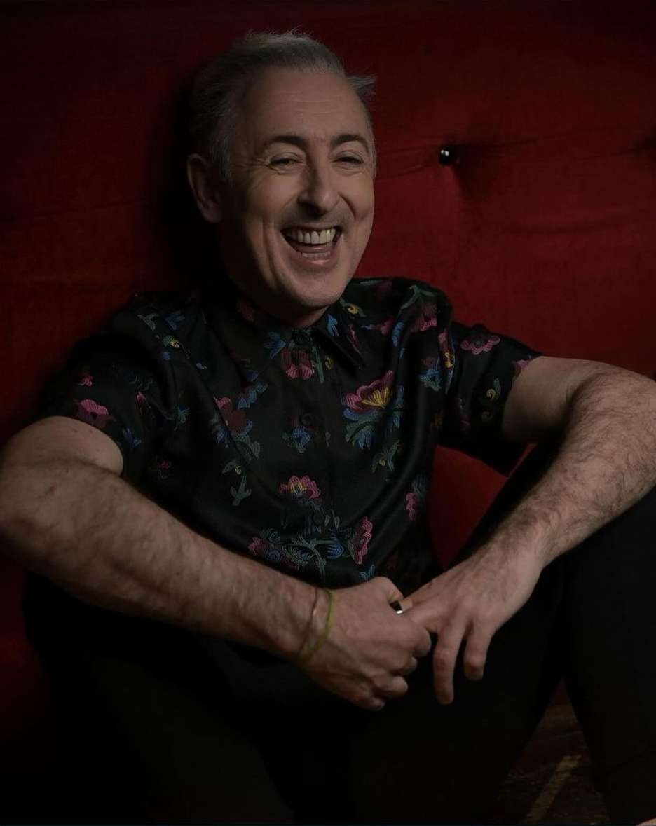 Portrait of smiling adult male, Alan Cumming, wearing a floral shirt, against a dark red background in an indoor setting with studio lighting.