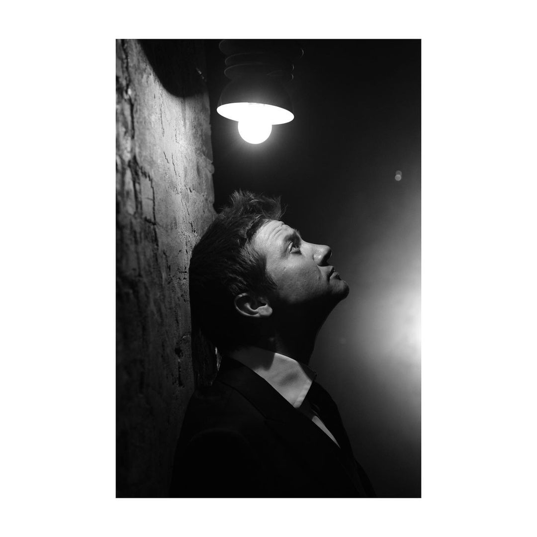 Monochrome portrait of a man in a suit looking up at a bright light, casting shadows against a textured wall, evoking a dramatic scene.