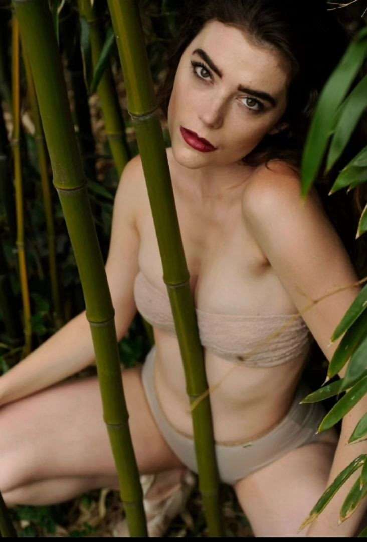 Adult female model in beige lingerie poses among bamboo stalks in outdoor natural setting, showcasing a portrait with natural lighting.