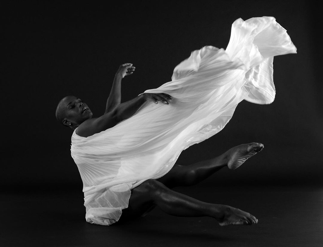 Black and white studio portrait of a Black woman in flowing white fabric, moving dynamically against a dark background. Fashion and dance.