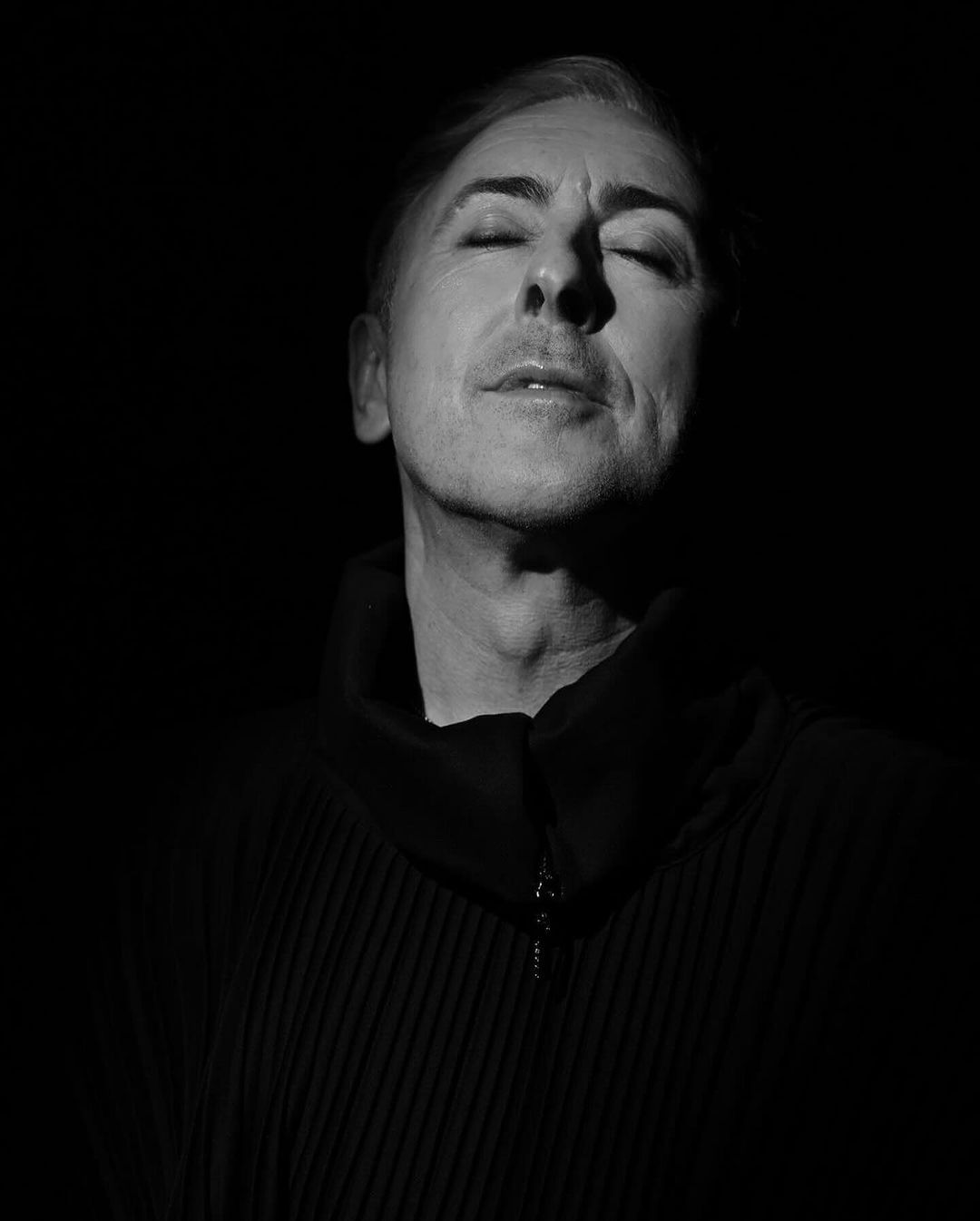 Monochrome portrait of a person with eyes closed, bathed in chiaroscuro lighting, wearing a ribbed top against a dark backdrop.