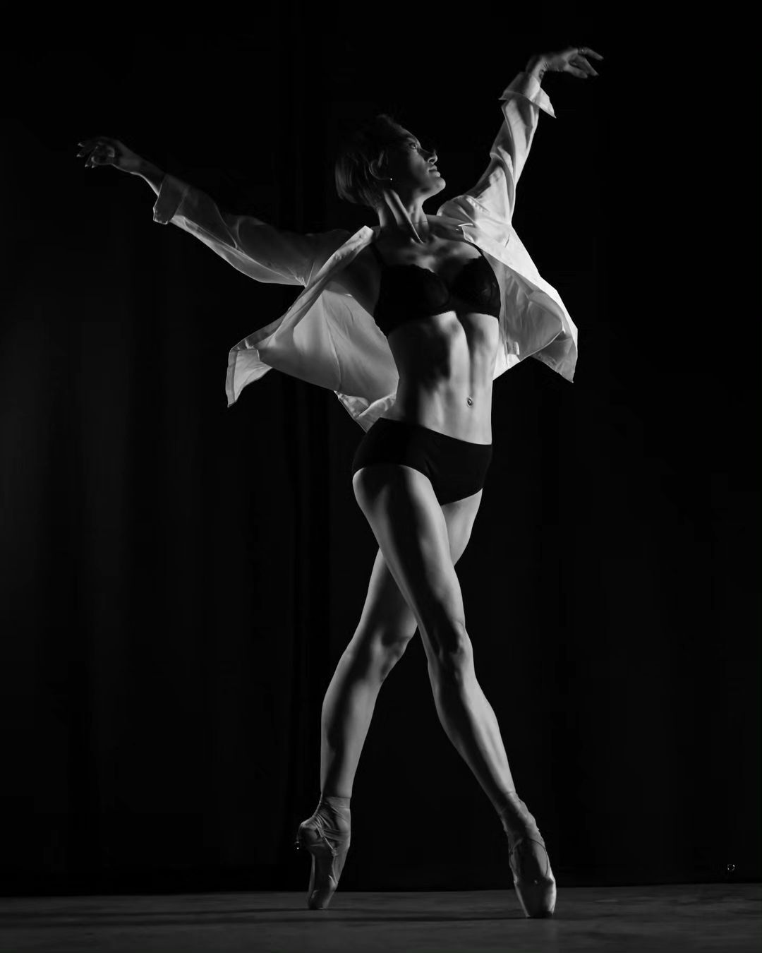 Black and white image of a ballet dancer in bra and shorts with an open white shirt, en pointe, with arms raised against a dark backdrop.