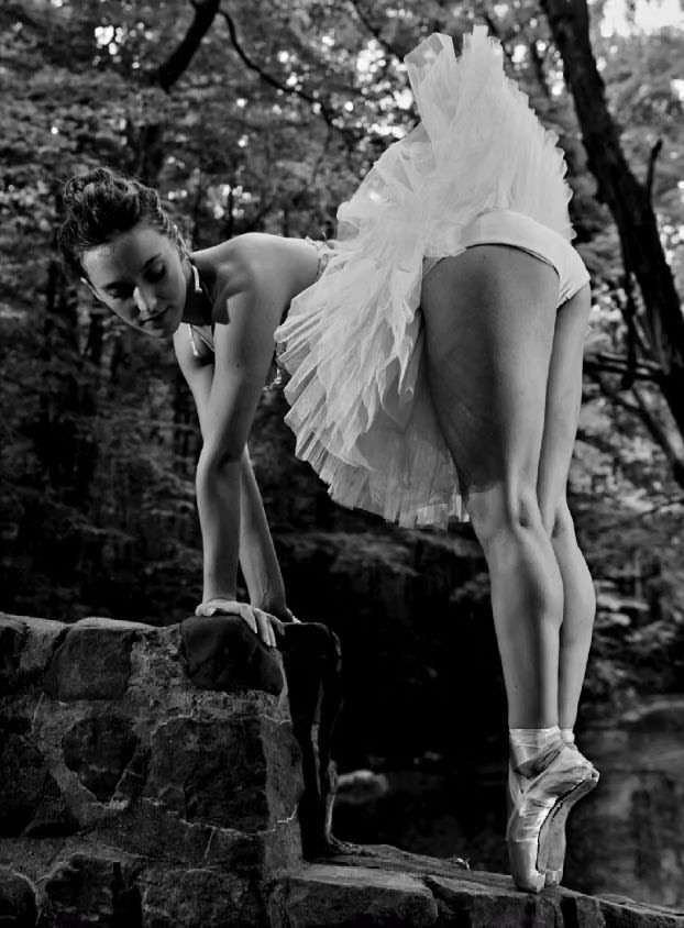 Black and white image of a ballet dancer in a tutu and pointe shoes on a stone wall, with a forest in the background.