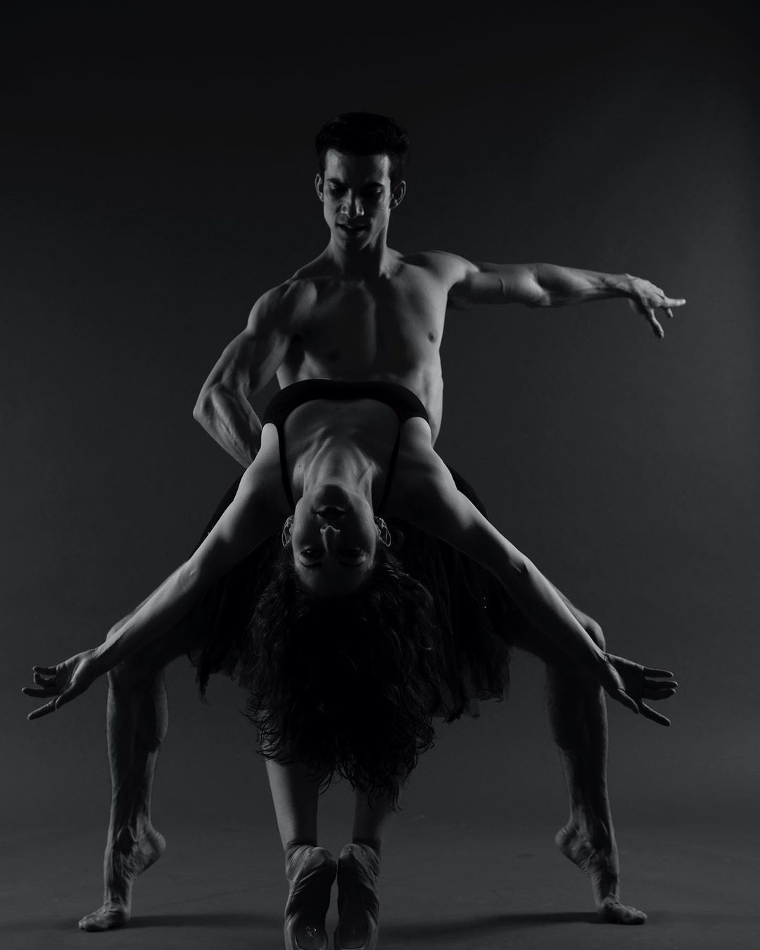 Dramatic black-and-white dance photo featuring a muscular male dancer supporting a woman upside down, lit to accentuate their forms.