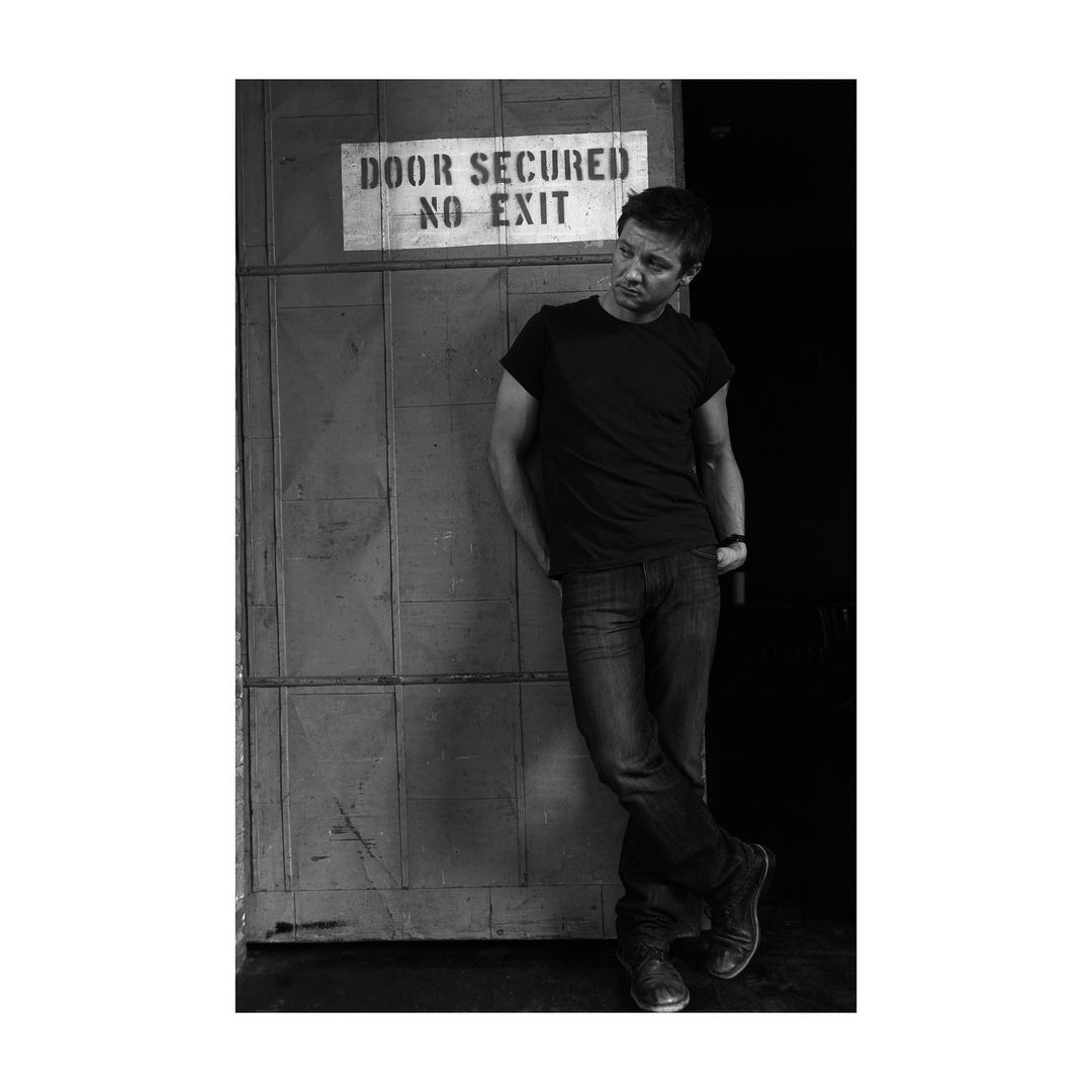 Black and white portrait of a man in a black t-shirt, leaning against a metal door with a sign reading 'Door Secured No Exit'.