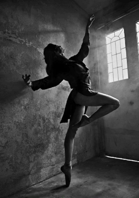 Black and white artistic image of a female ballet dancer en pointe against a textured wall in a room with sunlight streaming through windows.