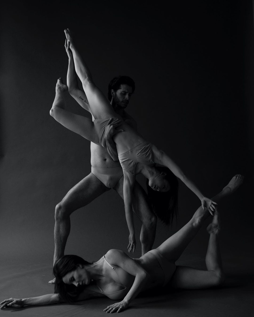 Monochrome studio shot of two women and one man in leotards striking dynamic poses, showcasing flexibility and strength in a dark grey setting.
