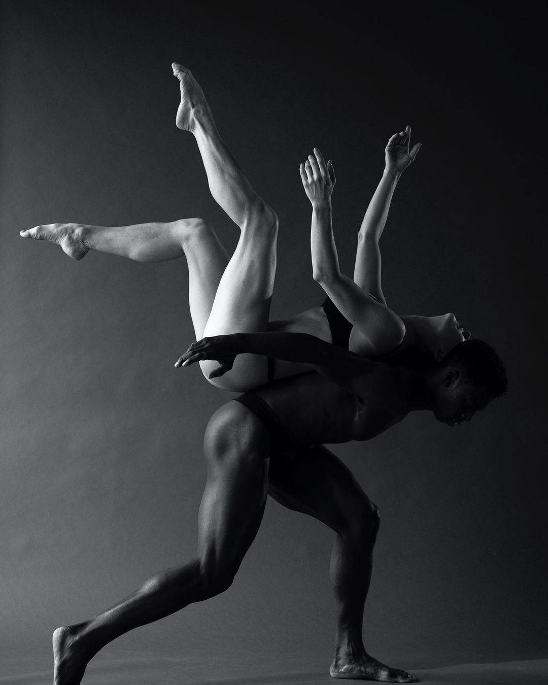 Monochromatic studio shot of two ballet dancers in dynamic poses, displaying strength and flexibility against a dark grey backdrop.