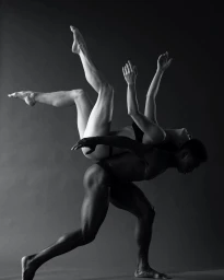 Black and white studio photograph of two dancers performing an athletic lift against a plain dark grey background.
