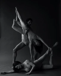 Monochrome studio photograph of three dancers performing a sculptural pose against a plain dark background.