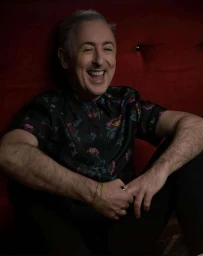 Portrait of Alan Cumming laughing while seated against a deep red tufted leather background, wearing a patterned short-sleeved shirt
