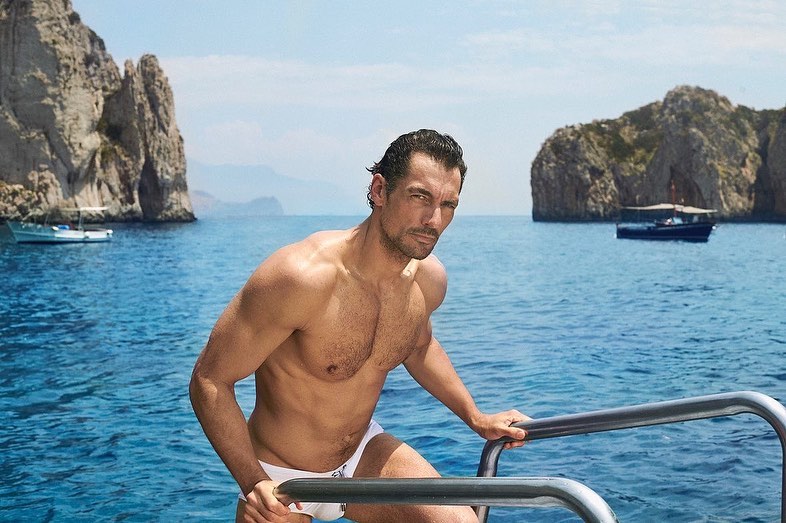 Adult male model on a boat in swimwear on the ocean in summer, set against a backdrop of rocky cliffs and blue sky.