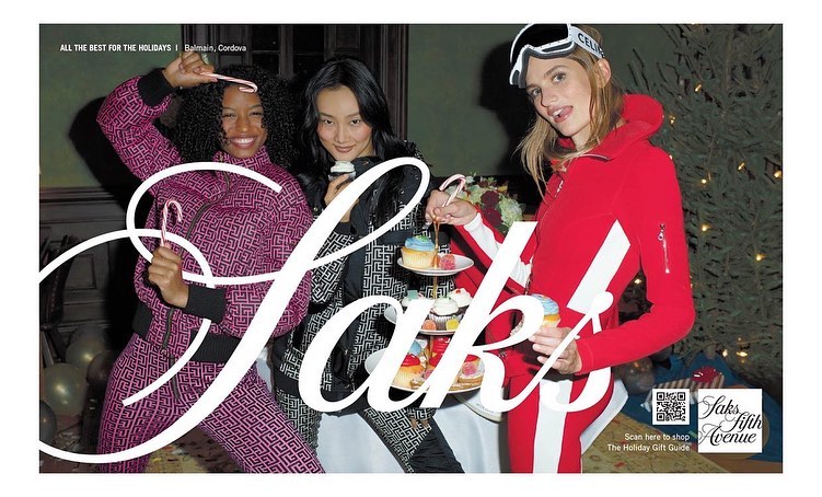 Three women in fashionable winter outfits pose indoors with cupcakes and candy canes in a holiday campaign ad for Saks Fifth Avenue.
