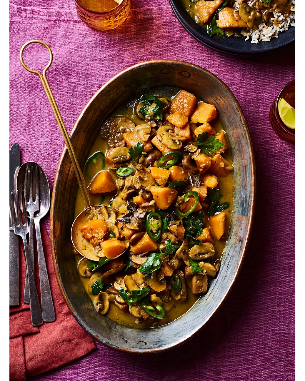 Overhead shot of a squash and mushroom curry dish in an oval copper serving dish with a ladle, set on a purple tablecloth.