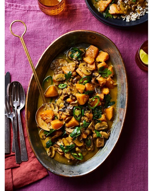 Overhead shot of a squash and mushroom curry dish in an oval copper serving dish with a ladle, set on a purple tablecloth.