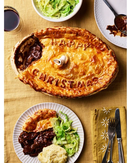 Overhead shot of a golden-crusted holiday pie with 'Happy Christmas' lettering, plated slice, side dishes, cutlery, and festive table setting.