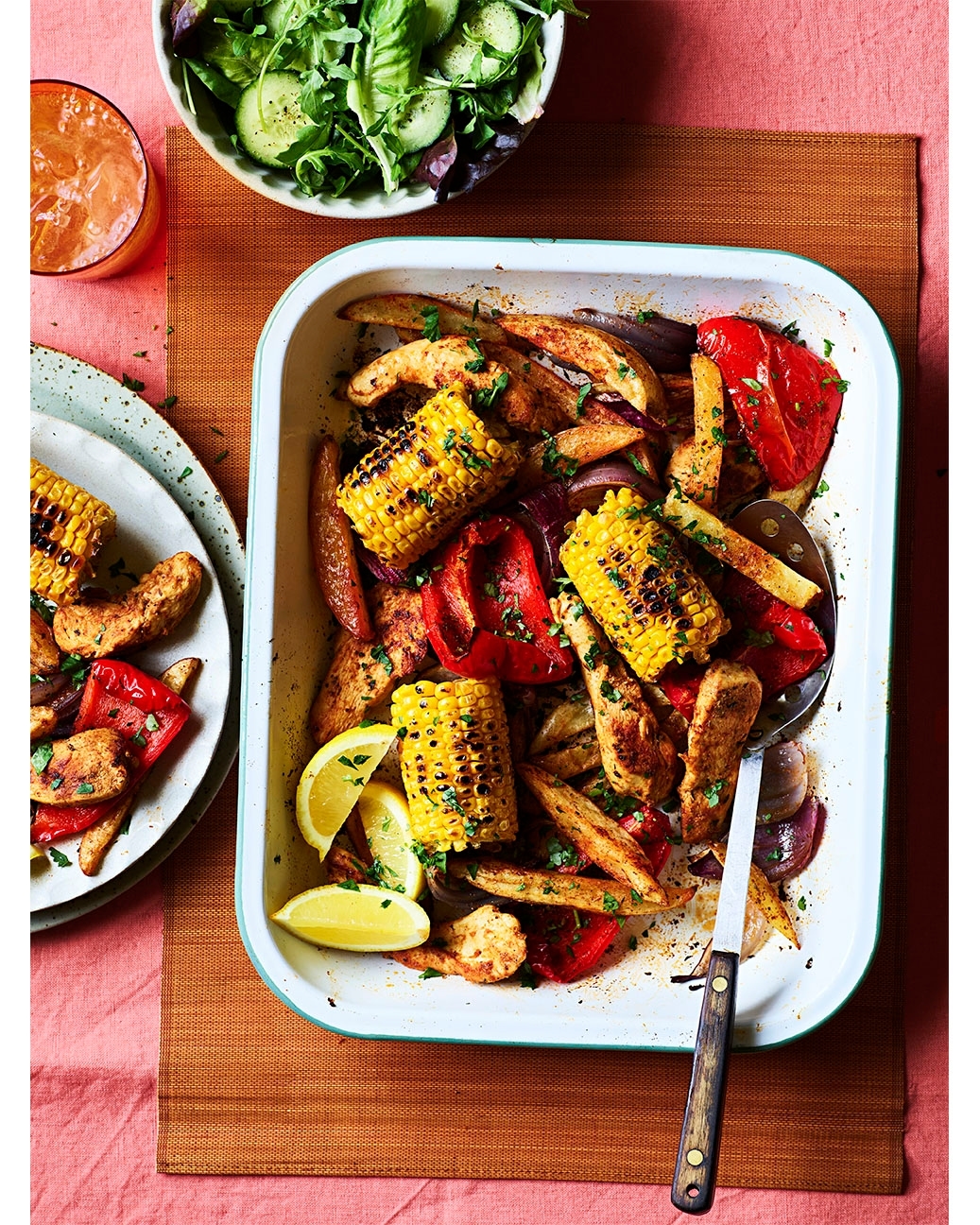 Overhead shot of grilled corn, red peppers, potatoes, red onions, and lemon slices arranged in a white baking pan on a woven mat.