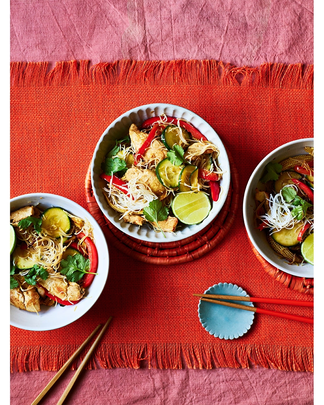 Overhead shot of three bowls of Asian-inspired noodle salad with vegetables, chicken, and lime, arranged on a red and pink tablecloth.