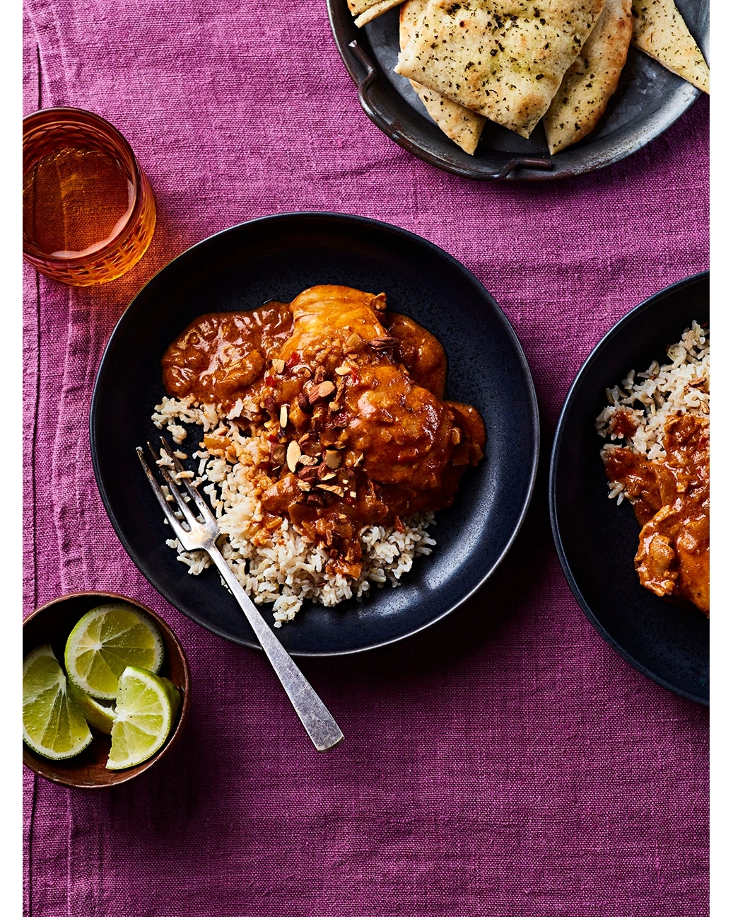 Overhead shot of Indian butter chicken with rice, lime wedges, and naan bread on a purple tablecloth.