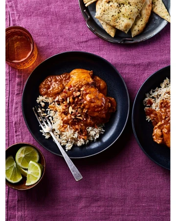 Overhead shot of Indian butter chicken with rice, lime wedges, and naan bread on a purple tablecloth.