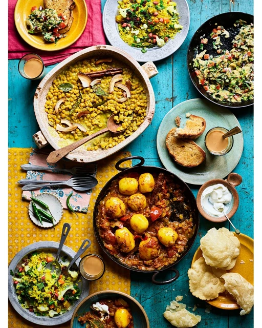 Overhead shot of a vibrant, textured table setting featuring an array of Indian dishes in bowls and pans, plus drinks and flatware.