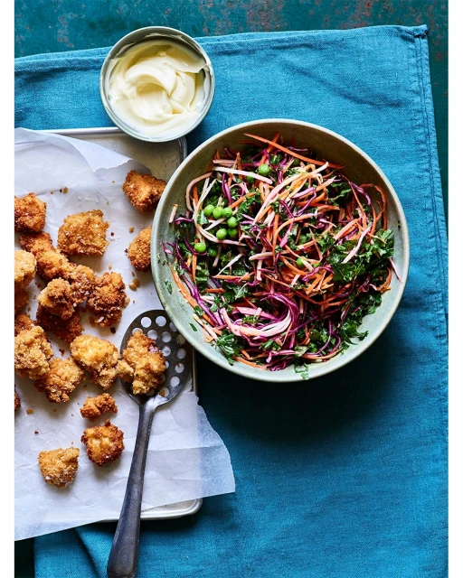 Overhead shot of fried chicken pieces, a colorful salad, and a bowl of sauce on a blue tablecloth. Food photography, studio lighting.