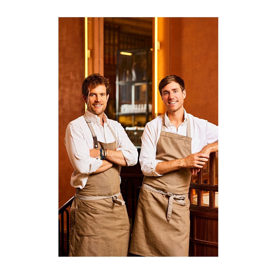 Two smiling chefs in aprons pose indoors, one with arms crossed, against an ornate orange wall and dark wood railing background.