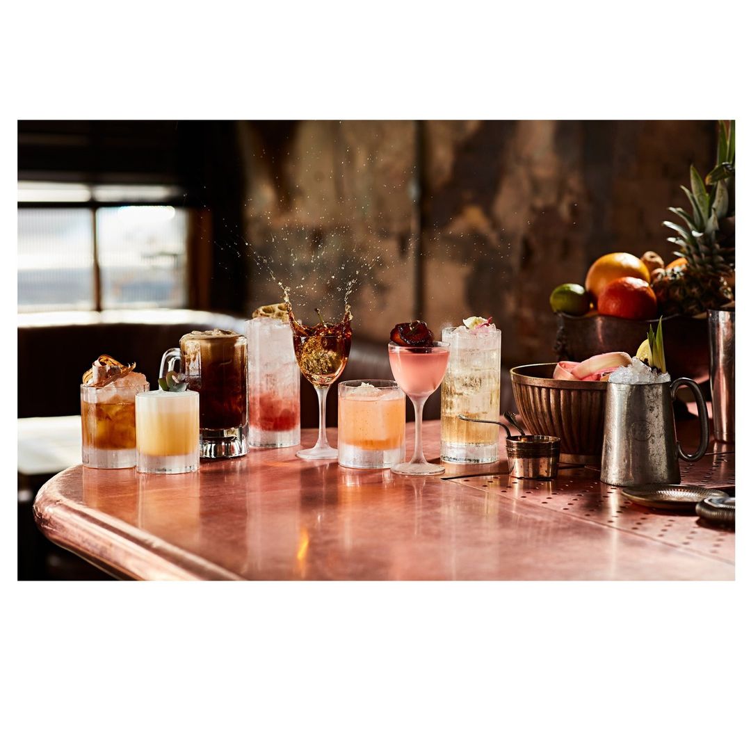 Several mixed cocktails in glasses of varying shapes and sizes are displayed on a copper bar top with fruit in background.