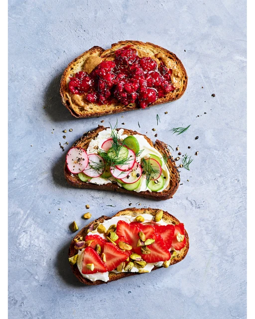 Three open-faced sandwiches on multigrain toast with toppings: raspberry jam and peanut butter, radish and cucumber, strawberry and pistachio.