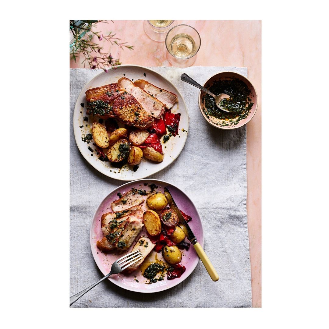 Overhead shot featuring two plates of sliced pork loin with roasted potatoes and red peppers, and a small bowl of green sauce.