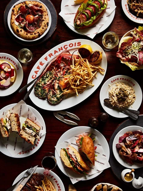 Overhead shot of a wood table covered in plates of food, including lobster, sandwiches, pizza, salad, and french fries.