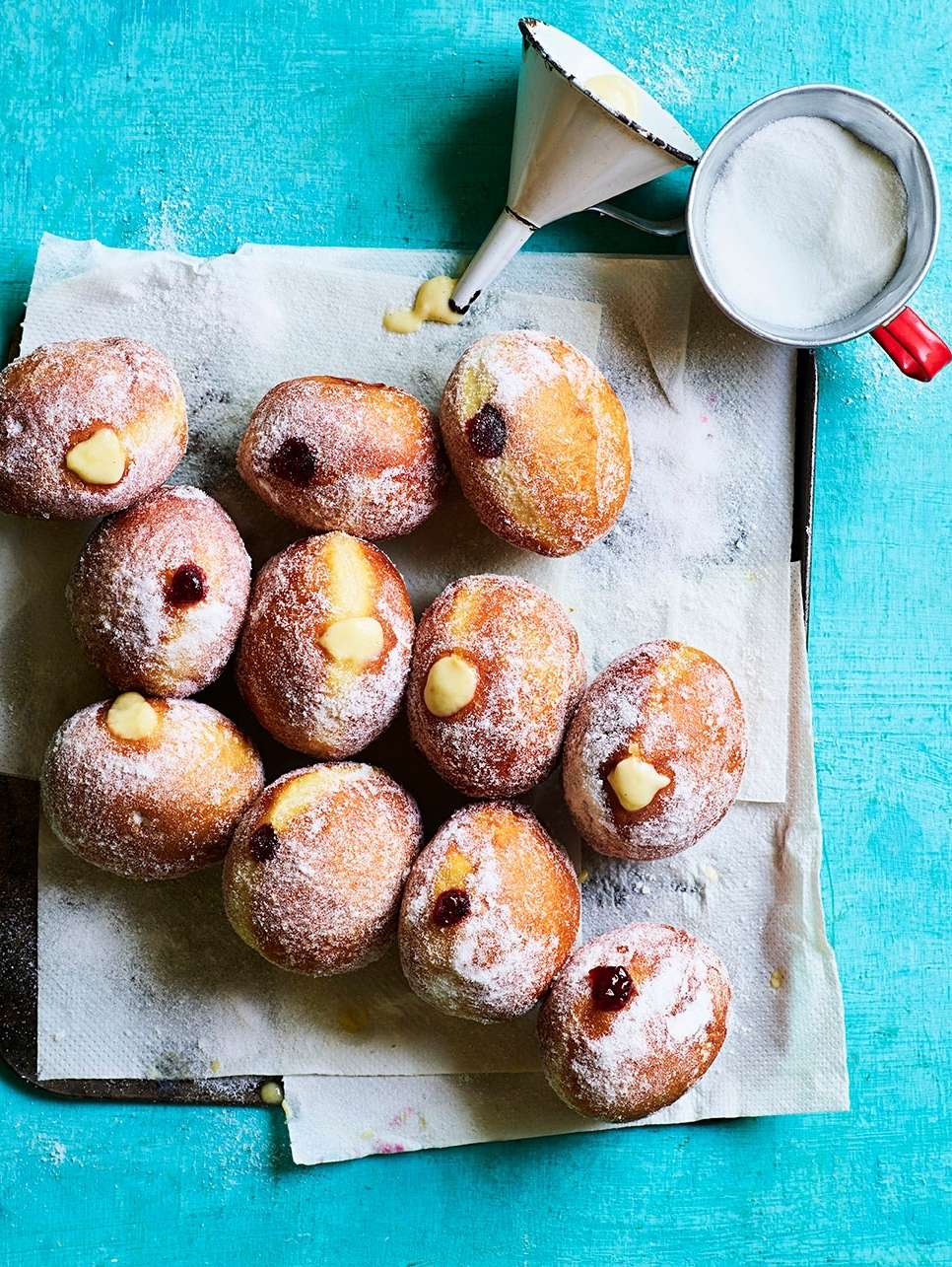 Overhead shot of a dozen cream and jam filled doughnuts covered in powdered sugar, arranged on parchment paper over a blue surface.