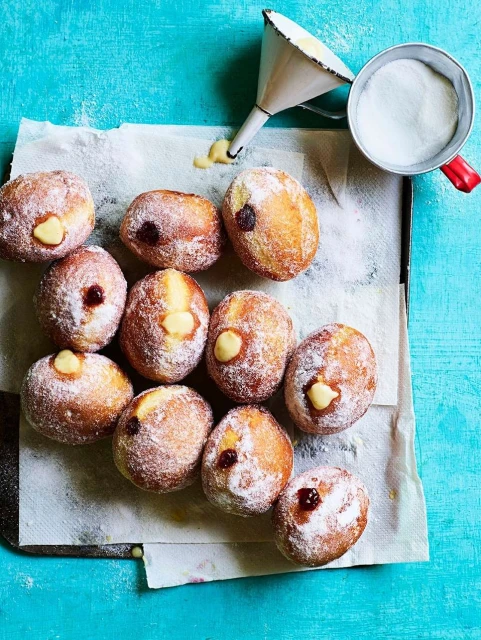Overhead shot of a dozen cream and jam filled doughnuts covered in powdered sugar, arranged on parchment paper over a blue surface.