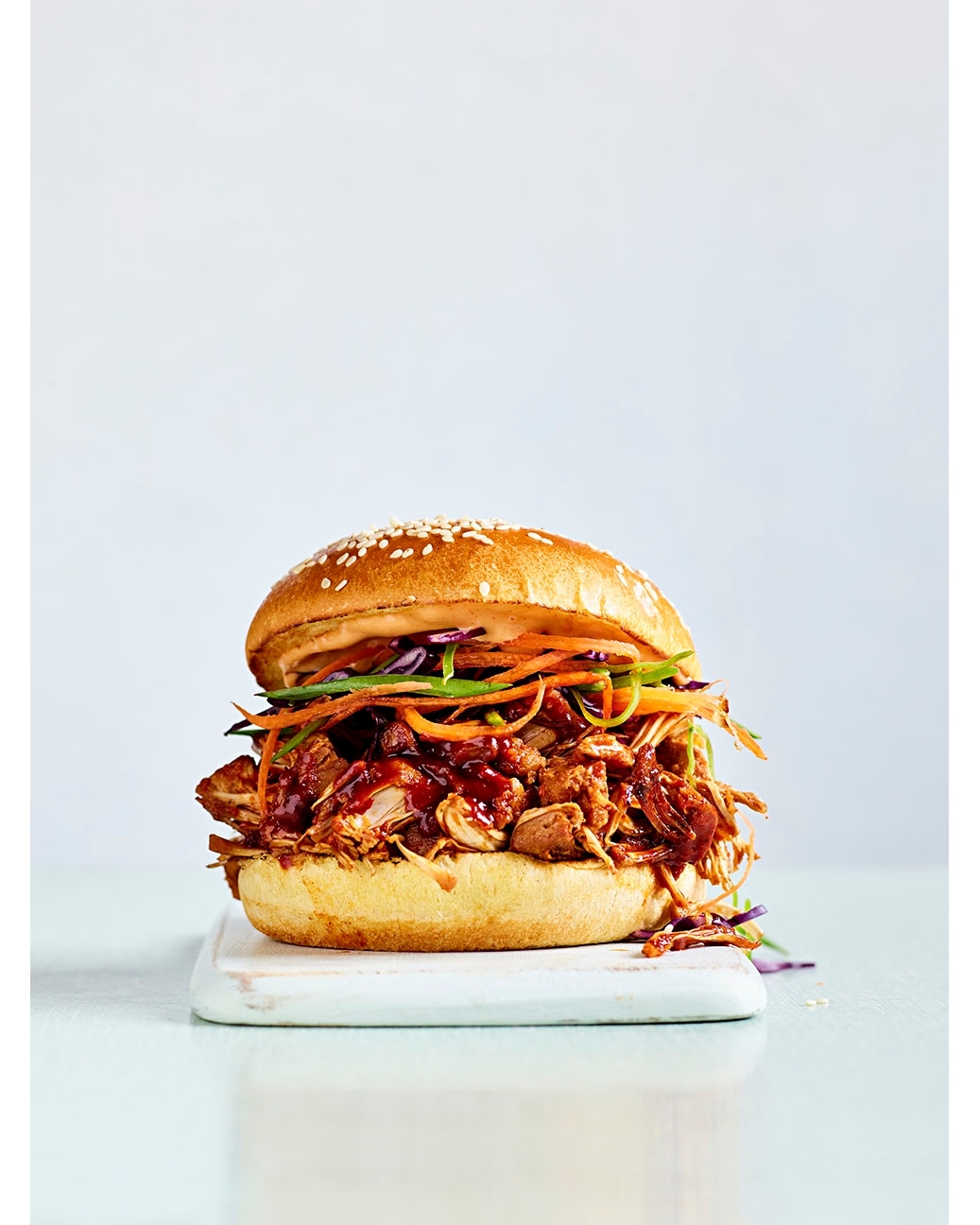 Close-up of a pulled pork sandwich with colorful slaw on a sesame seed bun, served on a white cutting board against a light background.