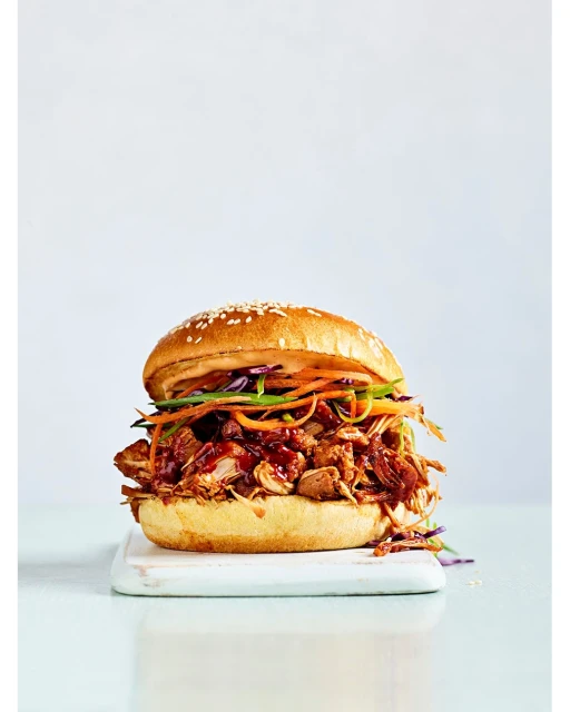 Close-up of a pulled pork sandwich with colorful slaw on a sesame seed bun, served on a white cutting board against a light background.