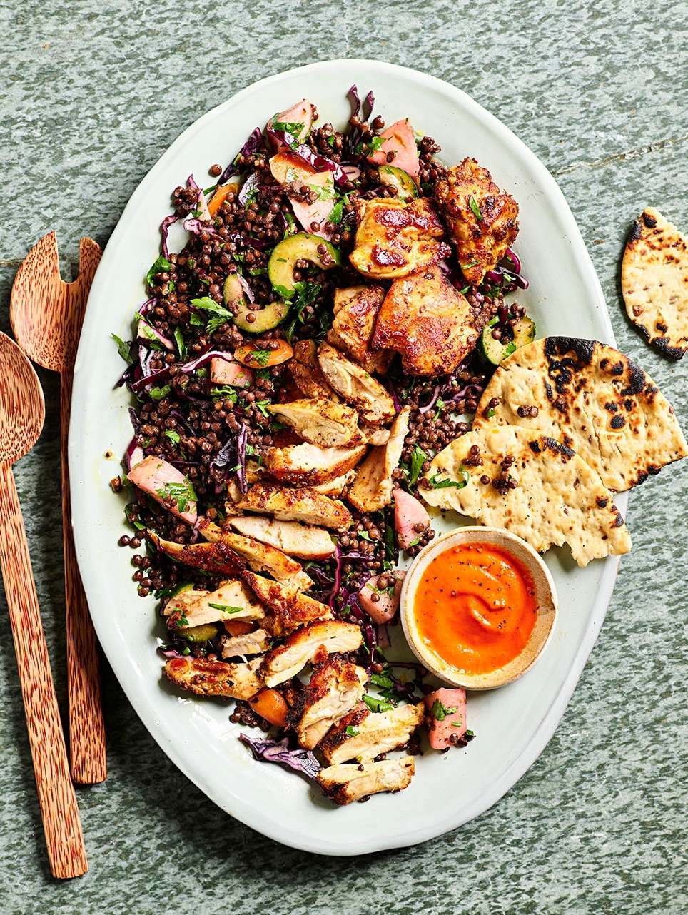 Overhead shot of a vibrant lentil salad with grilled chicken, flatbread, and a creamy orange sauce on a white oval platter.
