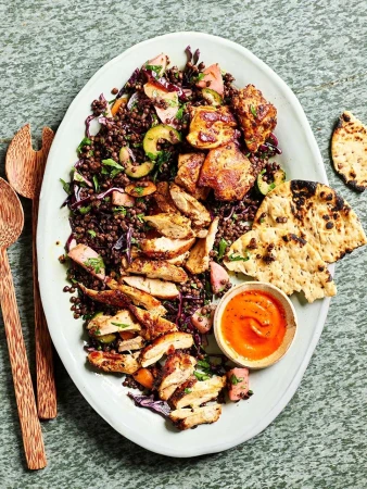 Overhead shot of a vibrant lentil salad with grilled chicken, flatbread, and a creamy orange sauce on a white oval platter.