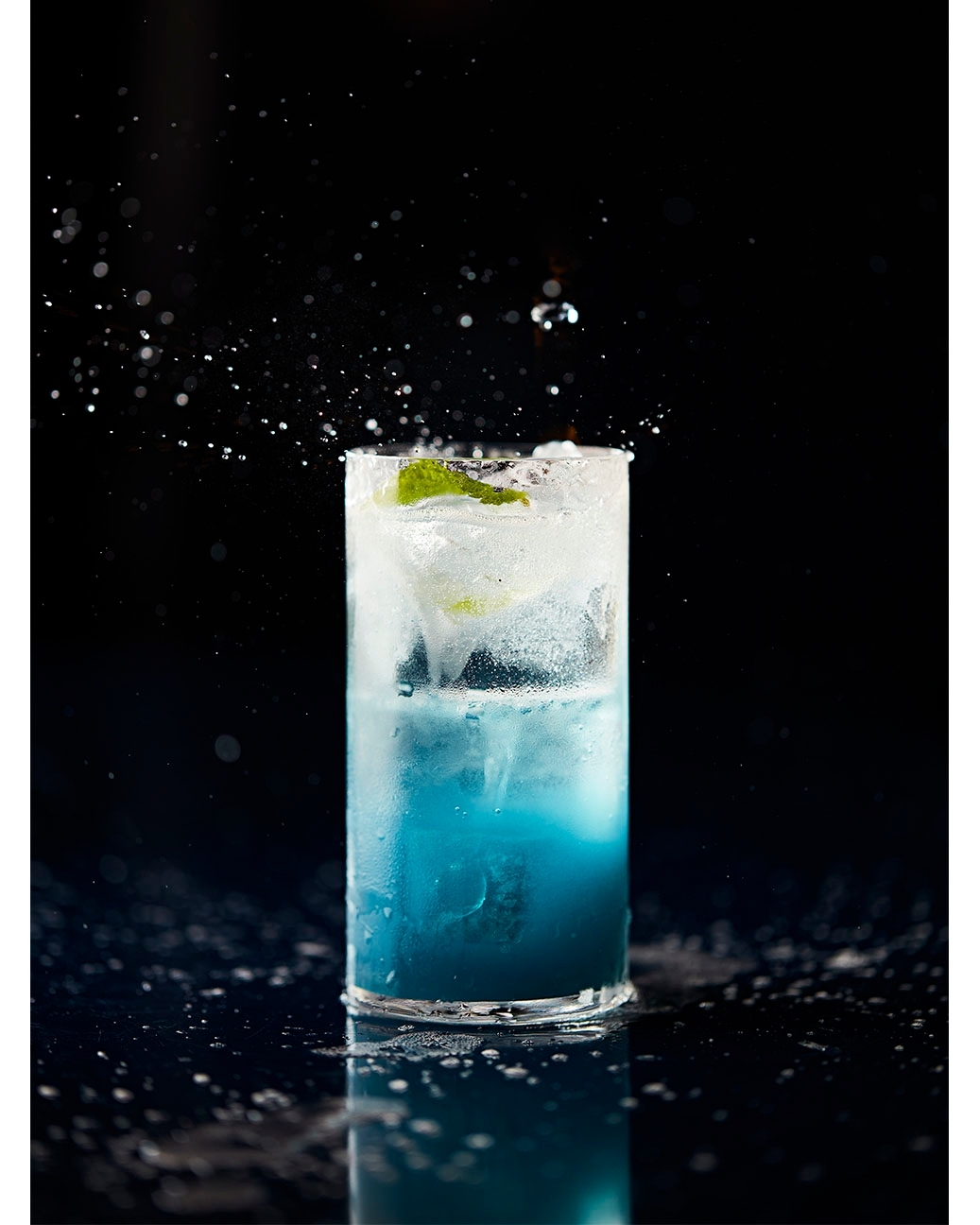 Tall glass filled with blue liquid and ice, garnished with a lime wedge, on a dark surface, with water droplets suspended in air.