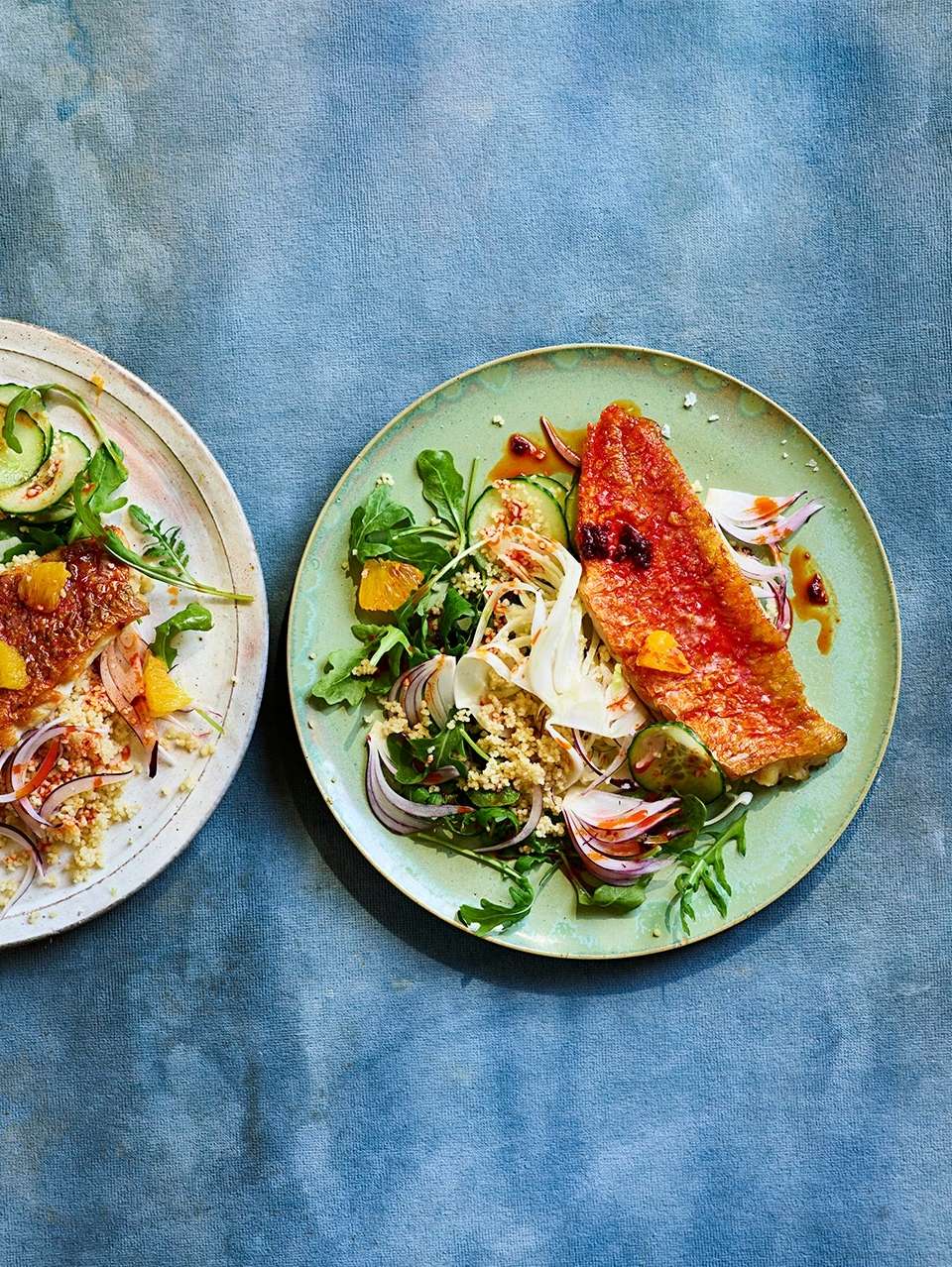 Overhead shot of two green plates featuring cooked red fish with greens, citrus, and couscous, set on a textured blue background.