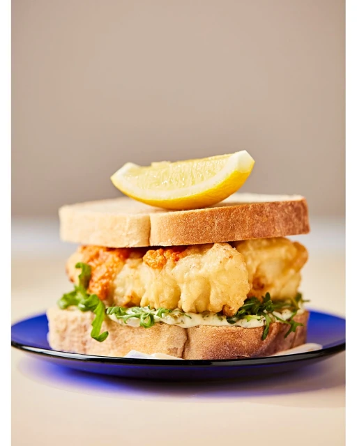 Overhead shot of a fish sandwich with lemon slice on a blue plate, featuring crispy fried fish, lettuce, tomato sauce, and white bread.
